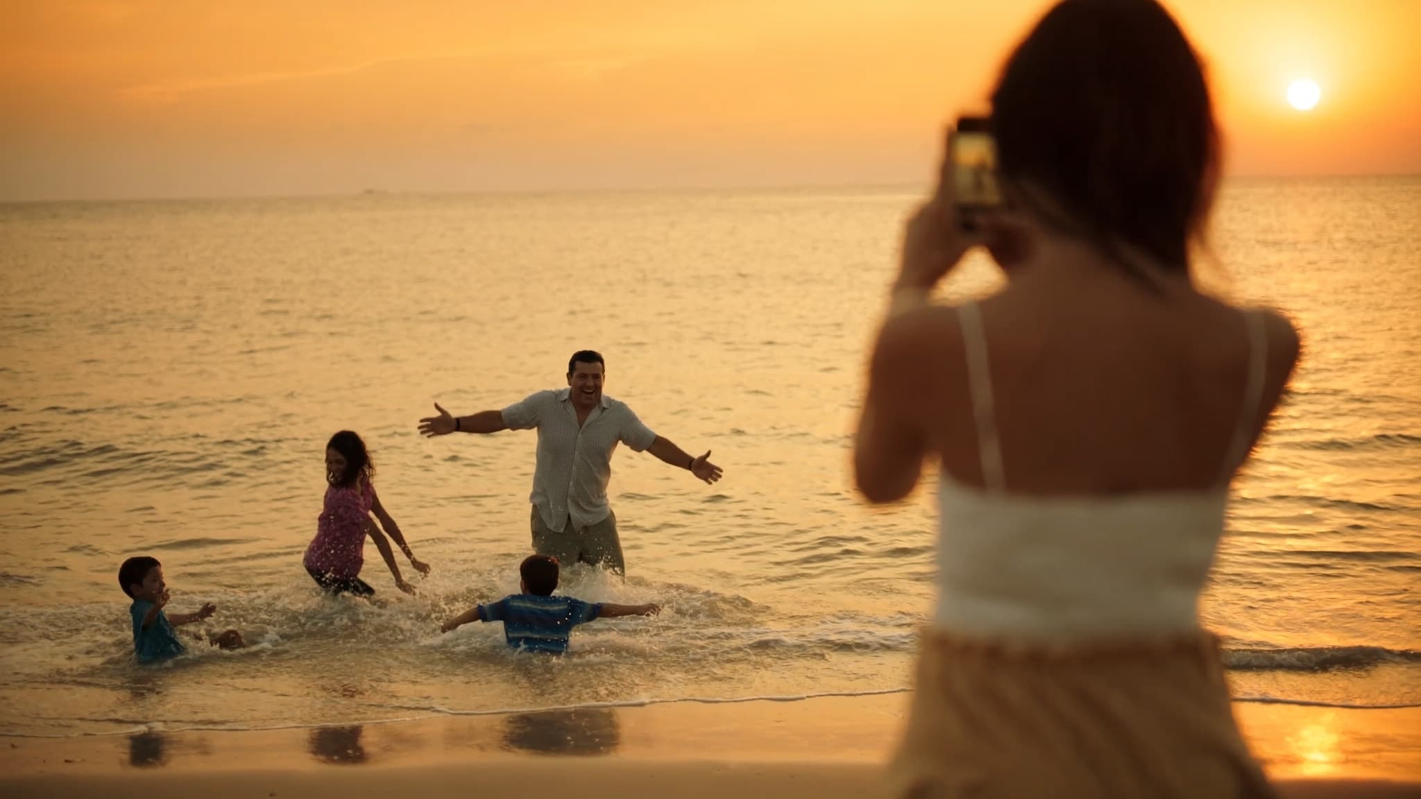 Mamá fotografiando a papá jugando con sus hijos en el mar al atardecer — seguro de vida