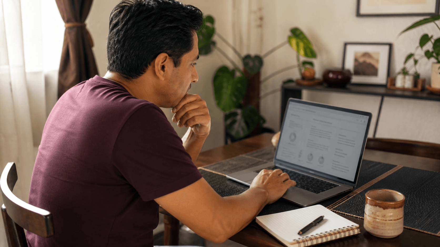 Hombre mexicano revisando opciones de inversión en su laptop, en la mesa del comedor de su casa, con una libreta y un café al lado.